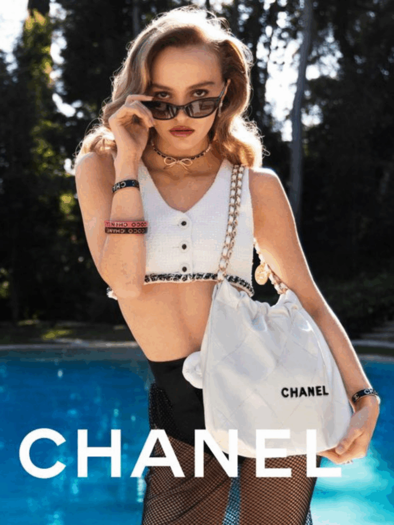 Lily Rose Depp stands outdoors in a backyard with trees in front of a pool, wearing a white Chanel crop top, black Chanel sunglasses, and a Chanel necklace. She holds her sunglasses slightly down on the bridge of her nose, looking directly at the camera, with a chic, confident pose.