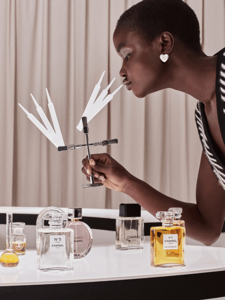 Black woman with a short, buzzed haircut smalling Chanel perfume test strips, holding a metal tool that is holding on to the strips. Chanel perfume bottles sit on a white table, and she wears a Chanel heart earring set against a muted pink background that is made up of curtains.
