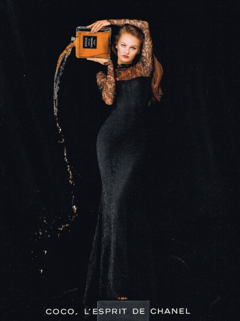 Vanessa Paradise stands against a deep black backdrop wearing a black lace-sleeved dress and red lipstick, holding a large Chanel perfume bottle. She is pouring out the perfume from the bottle, and it spills off to the side of her body. She poses barefoot with one arm over her head, creating a dramatic, luxurious sense.