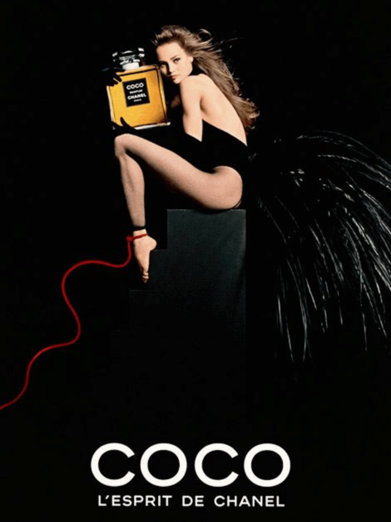 Vanessa Paradise sits on black stairs wearing a black bodysuit with black feathers like a bird tail, a high neckline and elbow-length black gloves, holding a large Coco Chanel perfume bottle. A red cord is around her ankle, implying she is not free to fly away. The studio lighting on her creates a contrast emphasizing the model, the perfume, and the bold, dramatic setting. The bottom of the image features the text "Coco L'esprit de Chanel."