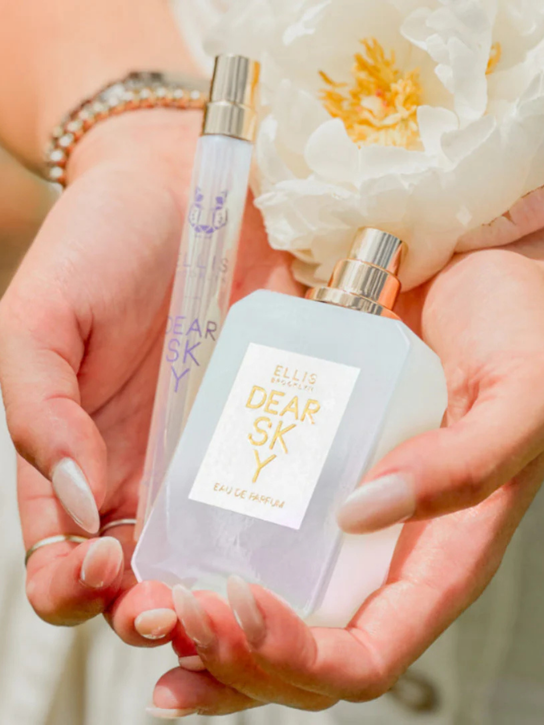 Full-size and travel-size bottles of Ellis Brooklyn Dear Sky perfume held in a woman's hands, showing the pink-to-blue ombré' bottle and the metallic travel spray. Her white iridescent nails, silver watch, and a small white flower on her wrist add a soft, feminine touch. The imagery highlights the fragrance's airy strawberry note and early-summer mood.