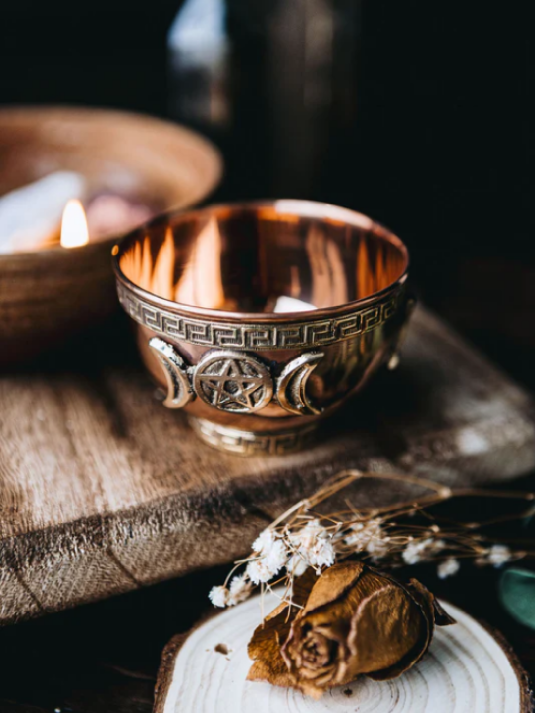 Copper-toned Triple Moon offering bowl displayed beside a wooden bowl with a lit candle inside it, dried blooms nearby with small white flowers on a wooden table. The setup evokes a strawberry-season altar aesthetic that complements Strawberry Moon offerings and early-summer ritual work.