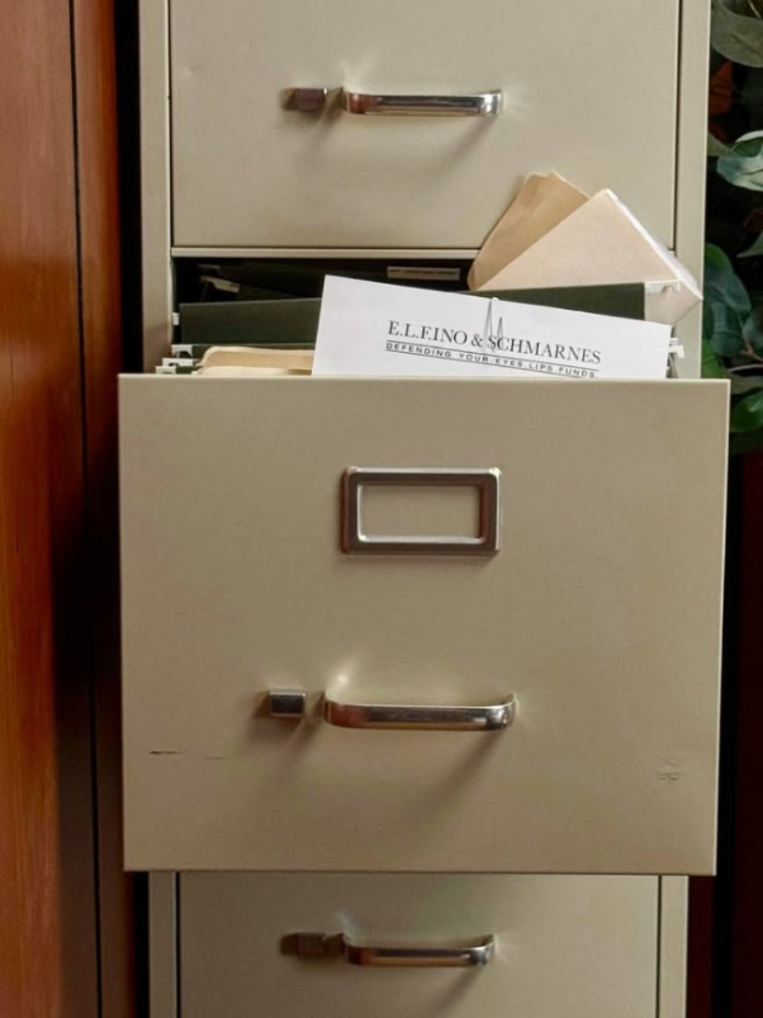 A filing cabinet with three drawers, where the middle drawer has a paper sticking out that reads "E.L.F.I.N.O & SCHMARNES, defending your eyes, lips, funds" used to illustrate the Matt Rife beauty controversy.