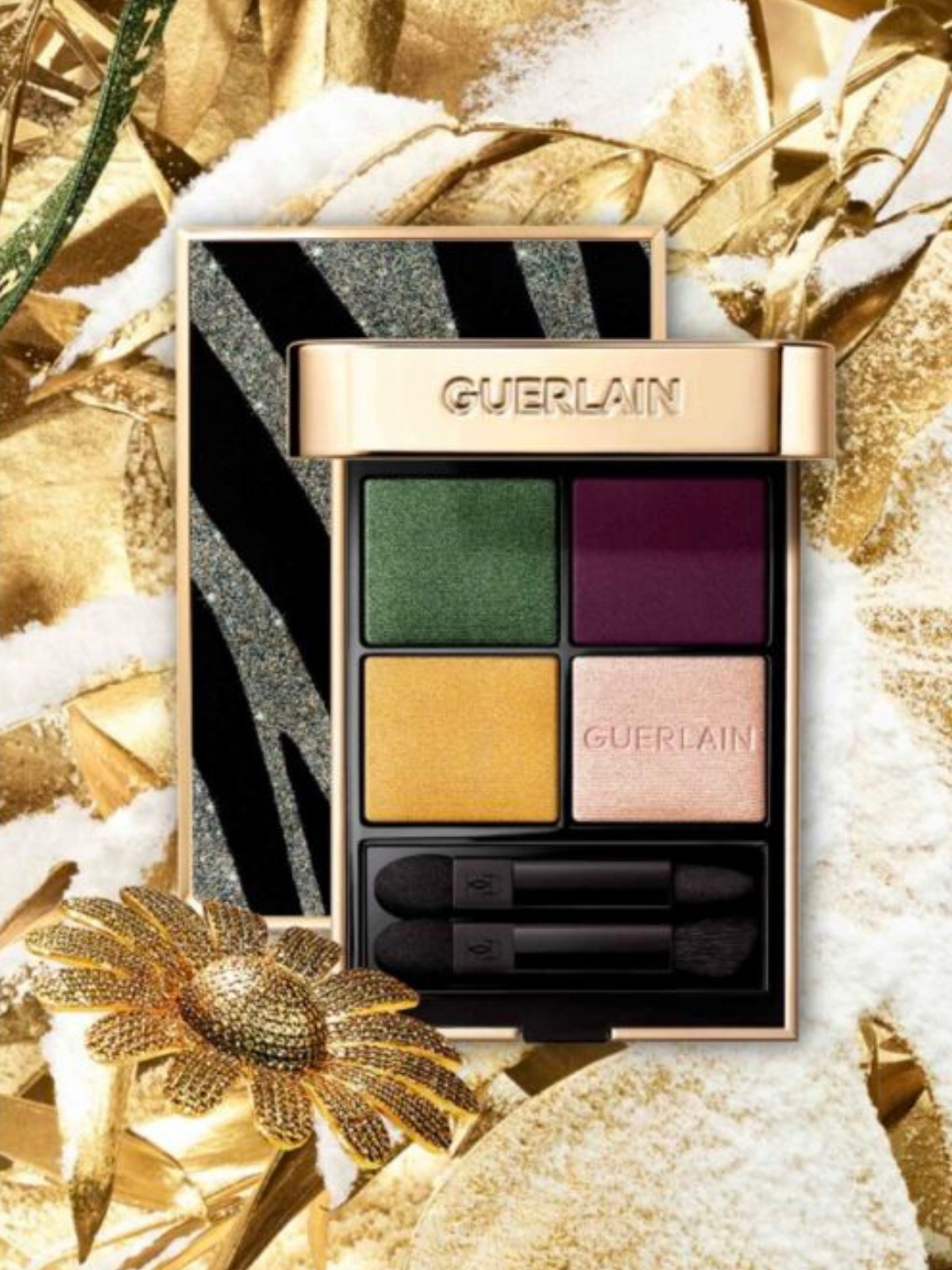 Guerlain G879 glittery Tiger eyeshadow palette from the 2023 Christmas collection featured in a holiday ad campaign with gold nature surrounding it while dusted in faux snow. One palette stands up closed and another is directly in front of it, open and showcasing the colorful eyeshadows inside.