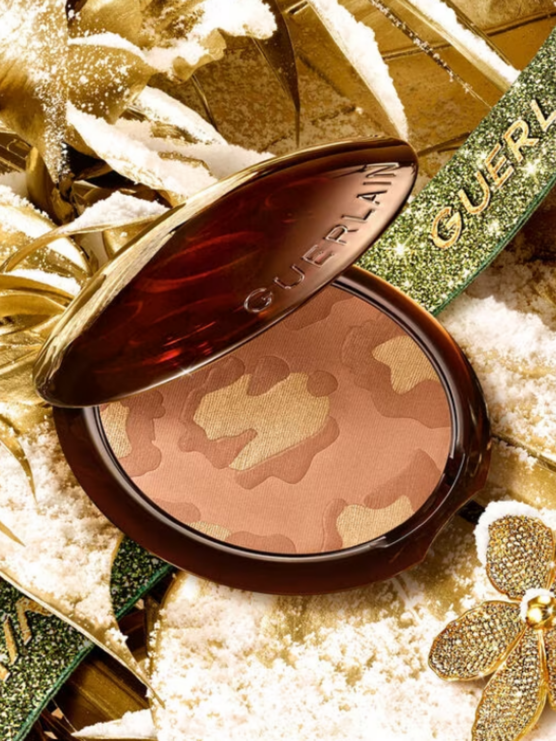 Guerlain Terracotta Gold Leopard Bronzer shown open in a holiday campaign setting, placed among gold leaves and floral elements with faux snow. The bronzer features a leopard-print pattern and is styled with a green ribbon displaying the Guerlain logo.