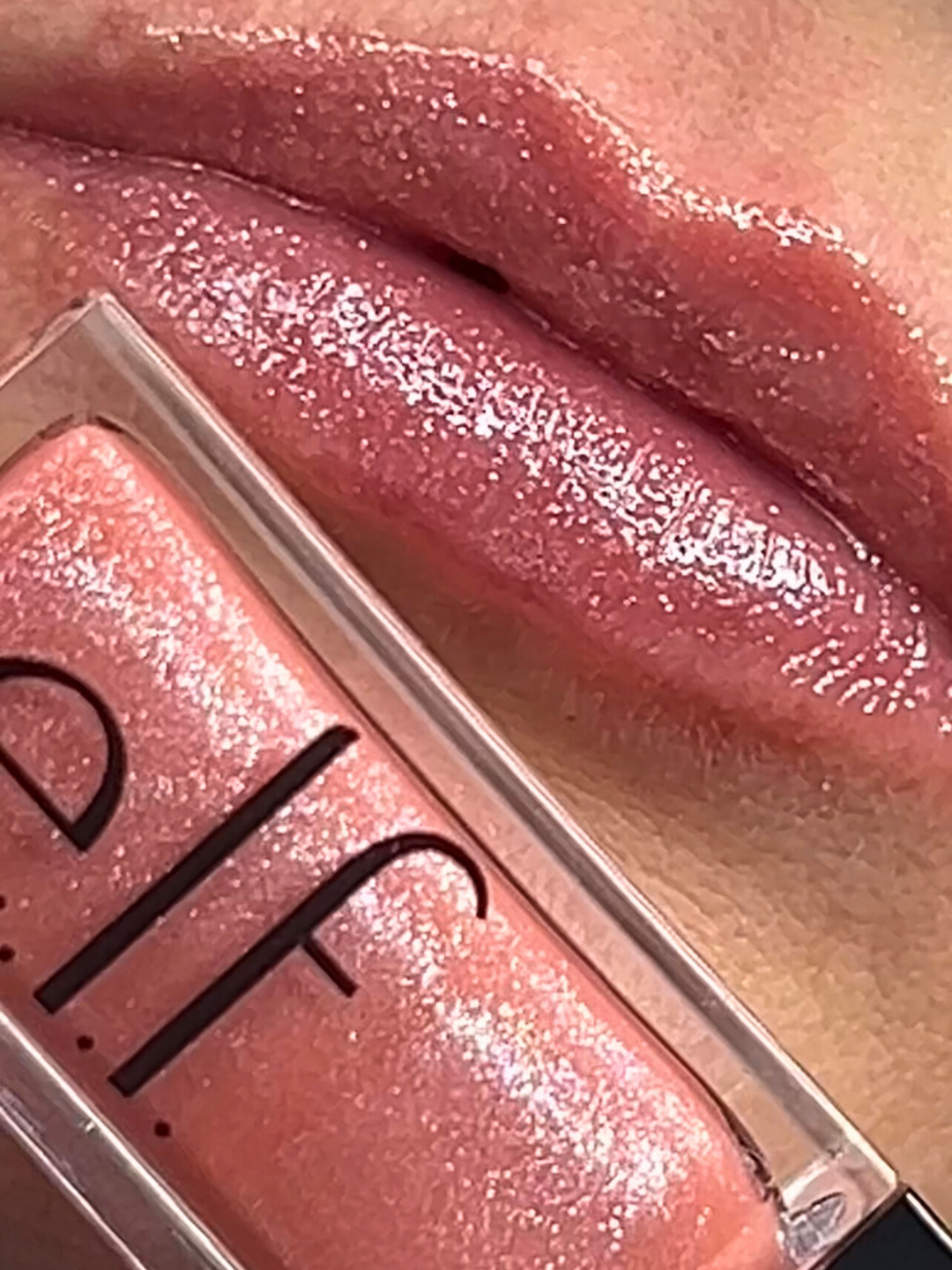 Detailed close-up of lips wearing e.l.f. Glow Reviver Lip Oil Glimmer in the shade "Princess Cut' with the product tube placed directly beneath the lips, highlighting the pink shimmer and glossy finish; beauty product swatch.