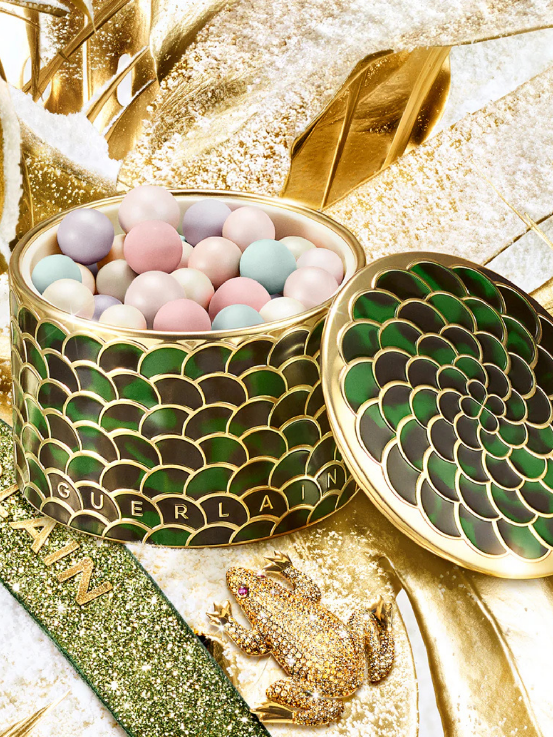 Guerlain Meteorites Fierce Glow Powder styled in a holiday campaign setting with gold nature-inspired decor, faux snow, and a green ribbon printed with the Guerlain logo. The open container reveals illuminating pearls beside a jeweled gold frog with pink eyes.