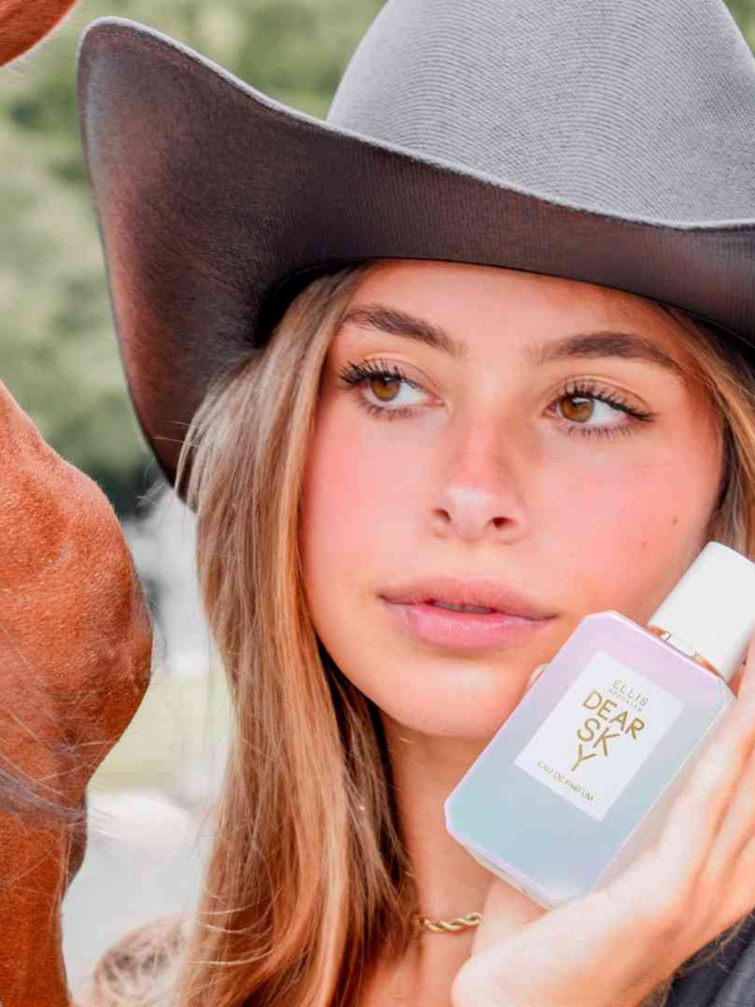Brunette woman standing beside a chestnut horse, holding the Ellis Brooklyn Dear Sky perfume bottle near her face. The rustic Western setting contrasts with the fragrance's soft strawberry note, creating a dreamy, Americana summer aesthetic.