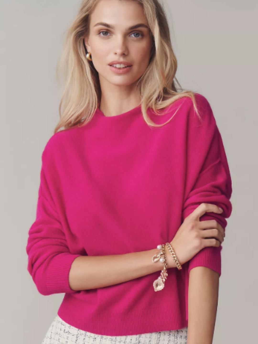 Woman wearing a pink raspberry cashmere mock-neck sweater by Pilcro, styled with gold earring, a charm bracelet, and a white-and -blue pleated skirt against a neutral backdrop.
