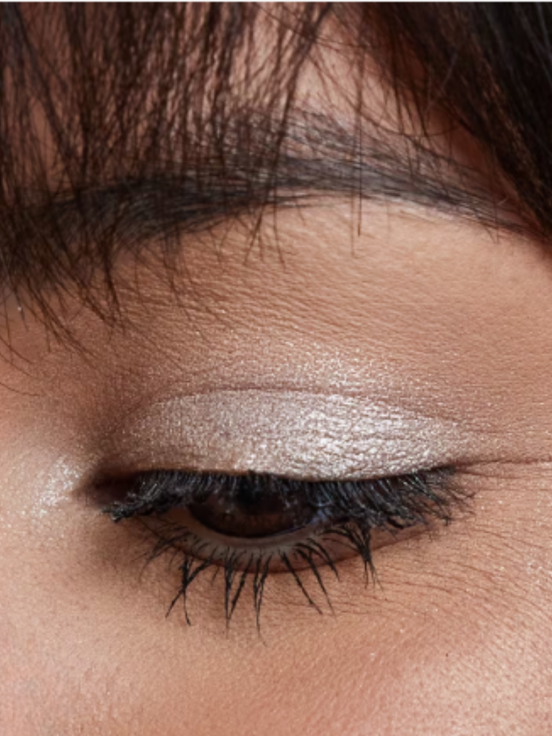 Medium-skin-tone model with dark hair wearing Urban Decay 24/7 Moondust Glitter Eyeshadow on her eyelid, eye half closed to show the shimmer and beauty effect.