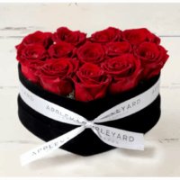 A black heart shaped box tied with a white Appleyard London ribbon, filled with upright preserved red roses designed to last a year, arranged as a luxurious floral gift that evokes elegance and self-care.