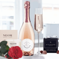 Images belong to Appleyard London A countertop display featuring a single preserved red rose from Appleyard London along NEOM Organics London candle, a light pink Adalina Prosecco bottle with chrome pink foil, a filled champagne flute, and a box of Appleyard London luxury truffle chocolates tied with a white ribbon- an elegant, indulgent arrangement that evokes a mood of celebration and self-care.