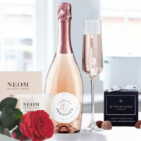 Images belong to Appleyard London A countertop display featuring a single preserved red rose from Appleyard London along NEOM Organics London candle, a light pink Adalina Prosecco bottle with chrome pink foil, a filled champagne flute, and a box of Appleyard London luxury truffle chocolates tied with a white ribbon- an elegant, indulgent arrangement that evokes a mood of celebration and self-care.
