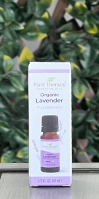 Front of Plant Therapy Organic Lavander Pure Essential Oil box in white packaging with purple trim, displayed outdoors on a table with lattice and ivy in the background. The box features a brown essential oil bottle with a black twist-off cap and a image of lavender flowers behind the bottle. The label reads "Plant Therapy Essential Oils Organic Lavender Pure Essential Oil."
