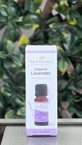 Front of Plant Therapy Organic Lavander Pure Essential Oil box in white packaging with purple trim, displayed outdoors on a table with lattice and ivy in the background. The box features a brown essential oil bottle with a black twist-off cap and a image of lavender flowers behind the bottle. The label reads "Plant Therapy Essential Oils Organic Lavender Pure Essential Oil."