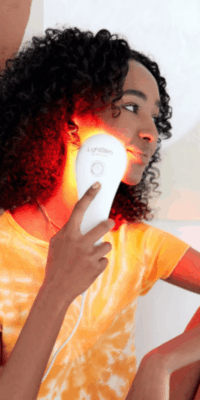Black woman with curly hair wearing an orange tie-dye shirt, holding a LightStim device against her cheek for red light therapy. She is seated with one elbow resting on her leg and the other hand holding the device, which is illuminated with red light.