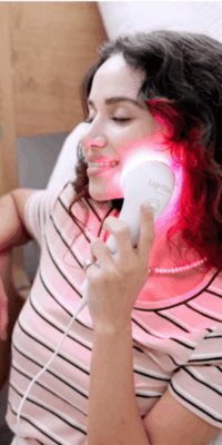 White woman with a slightly wavy brown hair, wearing a pink shirt with white and black stripes and a pearl necklace, seated with one elbow resting on a wooden surface. She is holding a LightStim device against her jawline, using red light therapy, and smiling with her eyes closed.