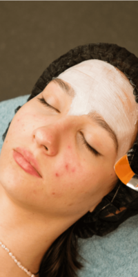 Young woman lying on a towel over a pillow with a brown headband and a pearl necklace, eyes closed, while an aesthetician applies a mask as a part of a post-acne extraction treatment. LightStim red light therapy can be used afterward to help reduce redness and promote faster healing.