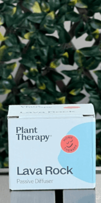 Front of Plant Therapy Lava Rock Passive Diffuser box in white packaging, displayed outdoors on a table with lattice and ivy in the background. The design features an irregular circular shape with a smaller circle inside that says "Home Therapy" with a smiley face illustration.