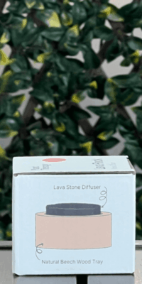 Side of Plant Therapy Lava Rock Passive Diffuser box in the white packaging, shown outdoors on an outdoor table with lattice and ivy in the background. The side panel features an illustration of the diffuser, showing a round lava stone resting in a circular natural beech wood tray. Labels read "Lava Stone Diffuser", with a squiggly arrow pointing to the lava stone, and "Natural Beech Wood Tray", with a circular arrow pointing to the wooden base.