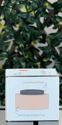 Side of Plant Therapy Lava Rock Passive Diffuser box in the white packaging, shown outdoors on an outdoor table with lattice and ivy in the background. The side panel features an illustration of the diffuser, showing a round lava stone resting in a circular natural beech wood tray. Labels read "Lava Stone Diffuser", with a squiggly arrow pointing to the lava stone, and "Natural Beech Wood Tray", with a circular arrow pointing to the wooden base.