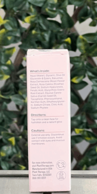 Back of Plant Therapy Rose with Bakuchiol Facial Serum box in light pink packaging, displayed outdoors on a table with lattice and ivy in the background. The back label lists ingredients, directions, cautions, and notes that the serum is vegan, packaged in the USA, and provides Plant Therapy's website, address, and phone number.