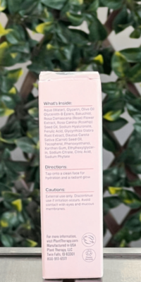 Back of Plant Therapy Rose with Bakuchiol Facial Serum box in light pink packaging, displayed outdoors on a table with lattice and ivy in the background. The back label lists ingredients, directions, cautions, and notes that the serum is vegan, packaged in the USA, and provides Plant Therapy's website, address, and phone number.