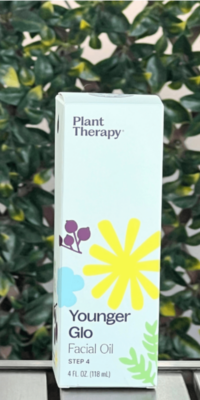 Front of Plant Therapy Younger Glo Facial Oil box in white packaging, displayed outdoors on a table with lattice and ivy in the background. The box features a purple, blue, and yellow flower with green leaves extending from the bottom corner, and text that reads "Younger Glo Facial Oil Step 4", with fluid ounces and milliliters, with the Plant Therapy logo at the top.