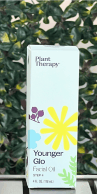 Front of Plant Therapy Younger Glo Facial Oil box in white packaging, displayed outdoors on a table with lattice and ivy in the background. The box features a purple, blue, and yellow flower with green leaves extending from the bottom corner, and text that reads "Younger Glo Facial Oil Step 4", with fluid ounces and milliliters, with the Plant Therapy logo at the top.