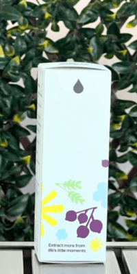 Side of Plant Therapy Younger Glo Facial Oil box in white packaging, displayed outdoors on a table with lattice and ivy in the background. The side panel features scattered purple, yellow, and blue flowers with green leaves, a small essential oil water drop icon at the top, and the phrase "Extract more from life's little moments" at the bottom.