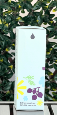 Side of Plant Therapy Younger Glo Facial Oil box in white packaging, displayed outdoors on a table with lattice and ivy in the background. The side panel features scattered purple, yellow, and blue flowers with green leaves, a small essential oil water drop icon at the top, and the phrase "Extract more from life's little moments" at the bottom.
