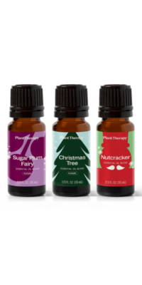 Three brown glass essential oil bottles with black caps from Plant Therapy Christmas Magic Essential Oil Blend 3 set. The first bottle, labeled "Christmas Tree", has a light blue snow-themed packaging with a green Christmas tree. The second, "Nutcracker", has a green label with a red nutcracker outline and white mustache. The third, "Sugar Plum", features a light purple label with a deep purple plum design.