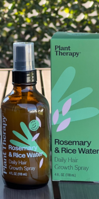 Plant Therapy Rosemary and Rice Water Daily Hair Growth Spray- a brown bottle with a black pump and clear cap, featuring a green leaf design split into three sections, three pink petals, and smiling green happy face labeled "Hair Therapy." The product sits beside its green box with matching design on an outdoor table with ivy-covered lattice in the background.