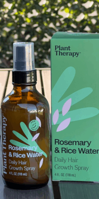 Plant Therapy Rosemary and Rice Water Daily Hair Growth Spray- a brown bottle with a black pump and clear cap, featuring a green leaf design split into three sections, three pink petals, and smiling green happy face labeled "Hair Therapy." The product sits beside its green box with matching design on an outdoor table with ivy-covered lattice in the background.