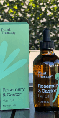 Plant Therapy Rosemary and Castor Hair Oil- a brown bottle with a black dropper cap and green "Hair Therapy" label showing a happy face, a green leaf parted into three sections, and pink petals. The product stands beside its matching green box on an outdoor table with ivy and lattice in the background.