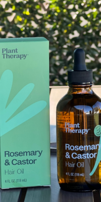 Plant Therapy Rosemary and Castor Hair Oil- a brown bottle with a black dropper cap and green "Hair Therapy" label showing a happy face, a green leaf parted into three sections, and pink petals. The product stands beside its matching green box on an outdoor table with ivy and lattice in the background.