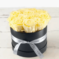 A round black box wrapped with a white Appleyard London ribbon sites on a cream faux-wood surface, filled with preserved yellow roses that last for a year, creating a bright, uplifting self-care gift.