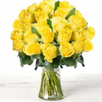 A clear glass vase filled with around 60 yellow roses sits on a cream faux-wood surface, creating a cheerful, uplifting floral arrangement that radiates warmth and self-care.