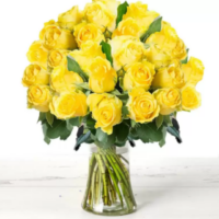A clear glass vase filled with around 60 yellow roses sits on a cream faux-wood surface, creating a cheerful, uplifting floral arrangement that radiates warmth and self-care.