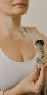 White woman wearing a white tank top with hair in a ponytail, holding a long, smoking sage bundle during a cleansing ritual. You can do this during your skincare routine using the Bakuchiol serum.