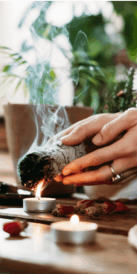 Woman holding a sage bundle over a small tea light candle that is on a coffee table. She wears a ring, with a houseplant and living room furnishings in the background. Bakuchiol is often incorporated in mindful skincare rituals alongside cleansing practices like sage smudging.