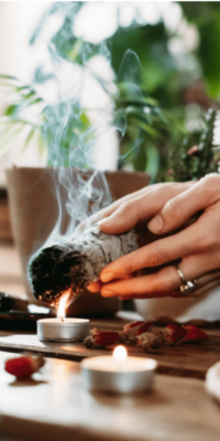 Woman holding a sage bundle over a small tea light candle that is on a coffee table. She wears a ring, with a houseplant and living room furnishings in the background. Bakuchiol is often incorporated in mindful skincare rituals alongside cleansing practices like sage smudging.