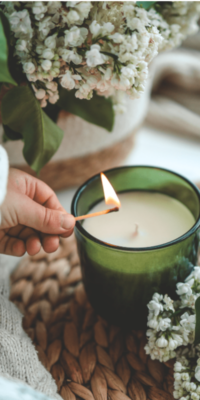 Green candle container with white candle wax inside being lit by a woman holding a match, placed on a woven wicker tray in a living room setting. Bakuchiol-infused skincare rituals often pair candlelight and calming practices to enhance mindfulness.