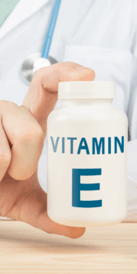 Close-up of a doctor's hand holding a white Vitamin E bottle with blue lettering, pointer finger on top and thumb at the bottom, highlighting the product. Bakuchiol is often paired with Vitamin E in skincare routines for enhanced benefits.