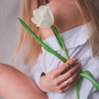 A white woman with blonde hair sits holding a single white tulip across her body and over her other arm, wearing gold rings on her ring and pointer fingers, capturing a quiet, reflective moment that embodies self-care and mindfulness.