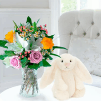 A round white table in a softly decorated dining space features a Jelly Cat stuffed animal-a white, long eared rabbit, and a glass vase filled with a mixed bouquet of pink roses, white lilies, yellow roses, small daisies, and delicate filler flowers from Appleyard London. Behind the table is a light blue-green padded chair with a silky floral pillow, creating an inviting, cozy scene that inspires moments of self-care.