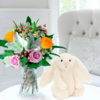 A round white table in a softly decorated dining space features a Jelly Cat stuffed animal-a white, long eared rabbit, and a glass vase filled with a mixed bouquet of pink roses, white lilies, yellow roses, small daisies, and delicate filler flowers from Appleyard London. Behind the table is a light blue-green padded chair with a silky floral pillow, creating an inviting, cozy scene that inspires moments of self-care.